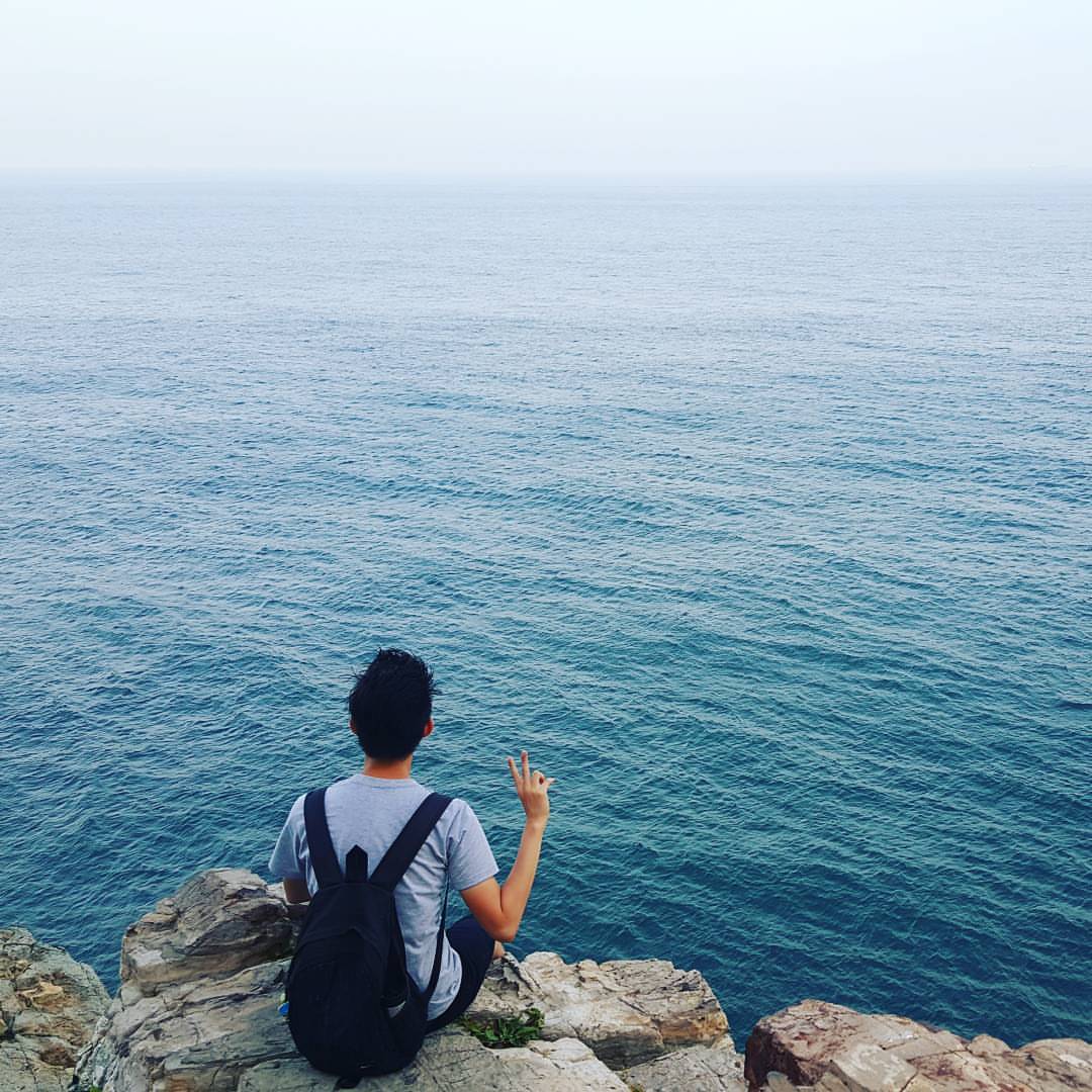 A photo from behind Endi&#39;s back, with Endi sitting on a rock cliff, facing the vast sea. His right hand forms a V pose.